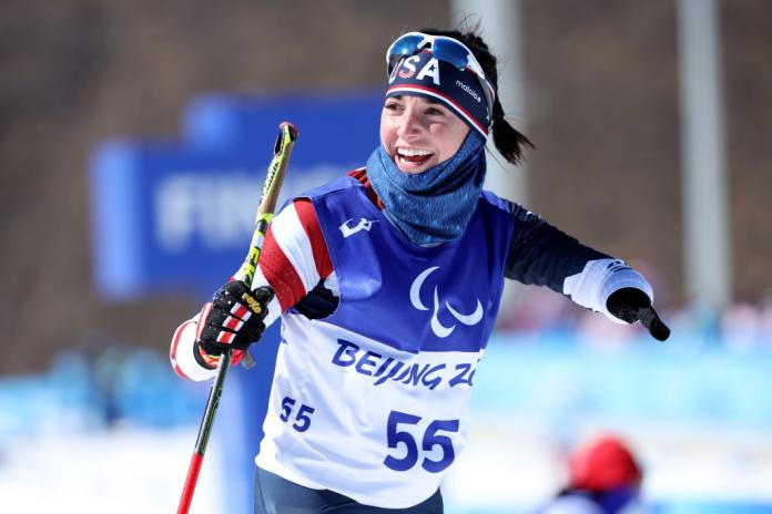 A female Para athlete wearing a USA skiing kit and a bib that says Beijing 2022 smiles after finishing her race